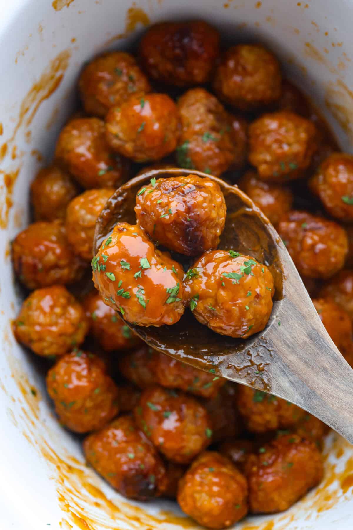Tender, juicy Slow Cooker Honey Buffalo Meatballs are simmered in the most tantalizing sweet heat sauce that everyone goes crazy for! Perfect appetizer or delicious, easy meal! Overhead shot of slow cooker honey buffalo meatballs with wooden spoon and three meatballs on it.