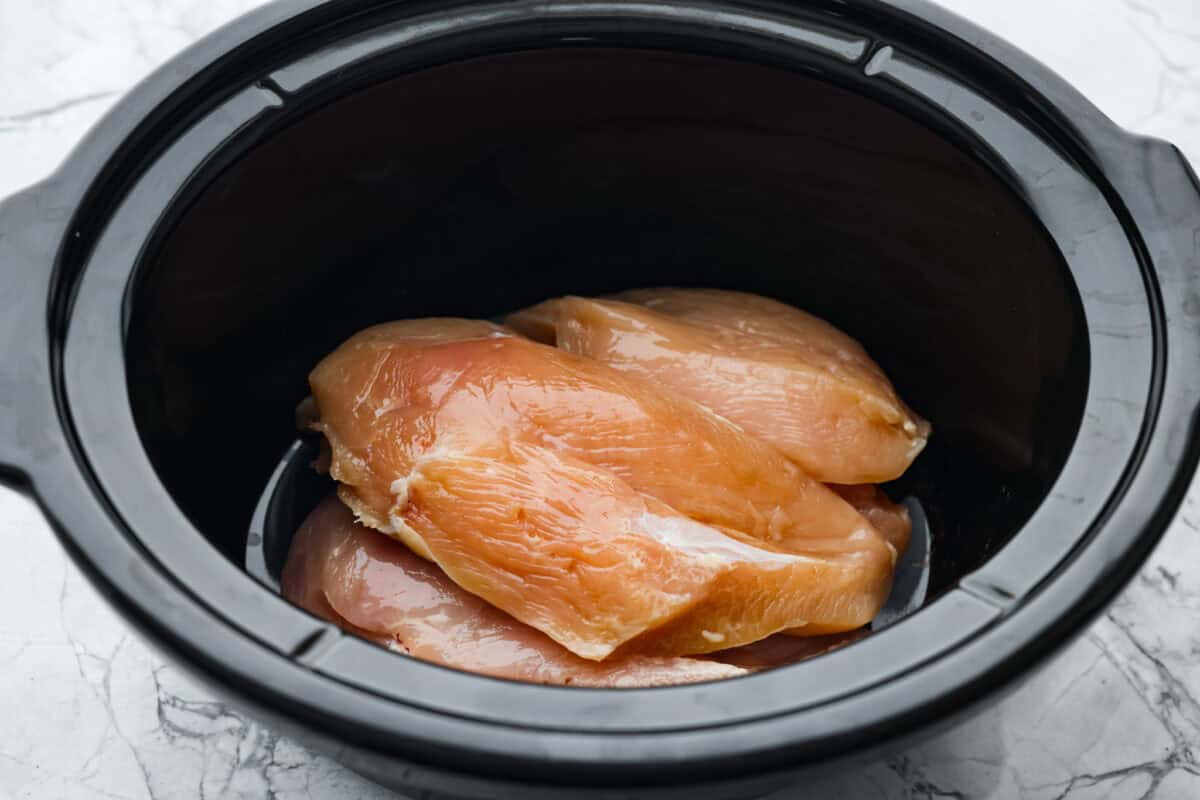 Slow cooker shredded BBQ chicken is a simple, zesty, and flavorful chicken. It can go in pasta dishes, over potatoes, in sandwiches, or in tortillas. This recipe is extremely versatile, tasty, and perfect for any time of year! Overhead shot of chicken breasts in teh bottom of the slow cooker.