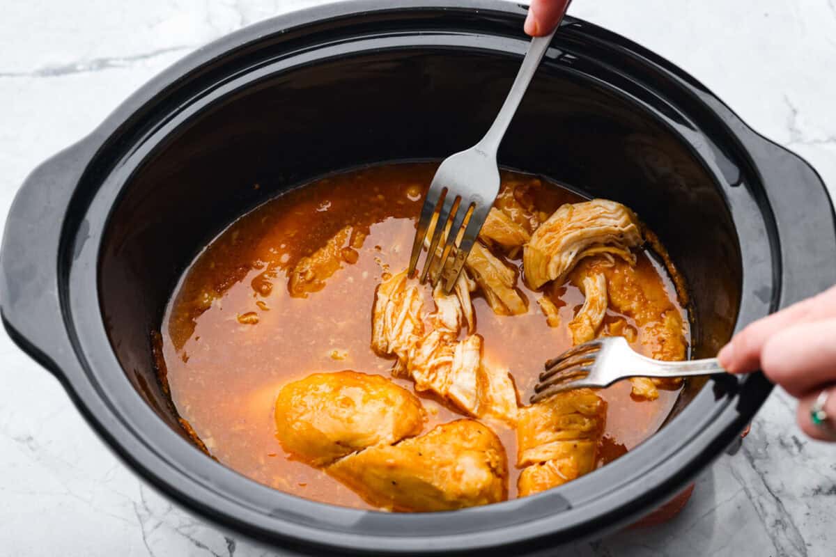 Slow cooker shredded BBQ chicken is a simple, zesty, and flavorful chicken. It can go in pasta dishes, over potatoes, in sandwiches, or in tortillas. This recipe is extremely versatile, tasty, and perfect for any time of year! Overhead shot of someone shredding the chicken with two forks in the slow cooker.