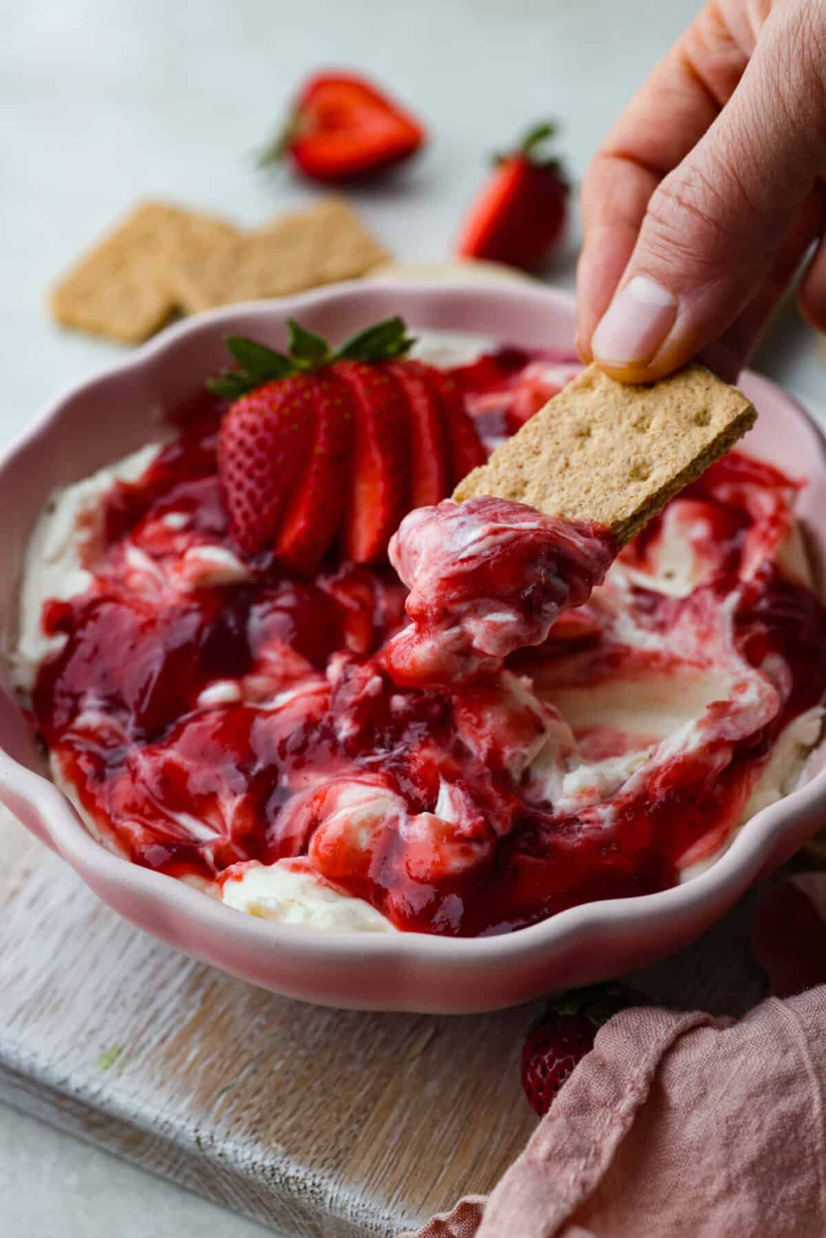 Strawberry cheesecake dip is an irresistible treat that tastes just like the classic but in a fun dip form! Whip it up in just 5 minutes! It's so good with graham crackers, fresh fruit, and cookies for a sweet, crowd-pleasing snack! Angle shot of someone dipping a graham cracker into the strawberry cheesecake dip.