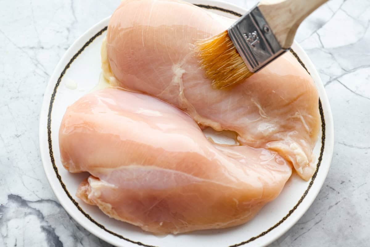 Someone brushing olive oil onto the two raw chicken breasts. 