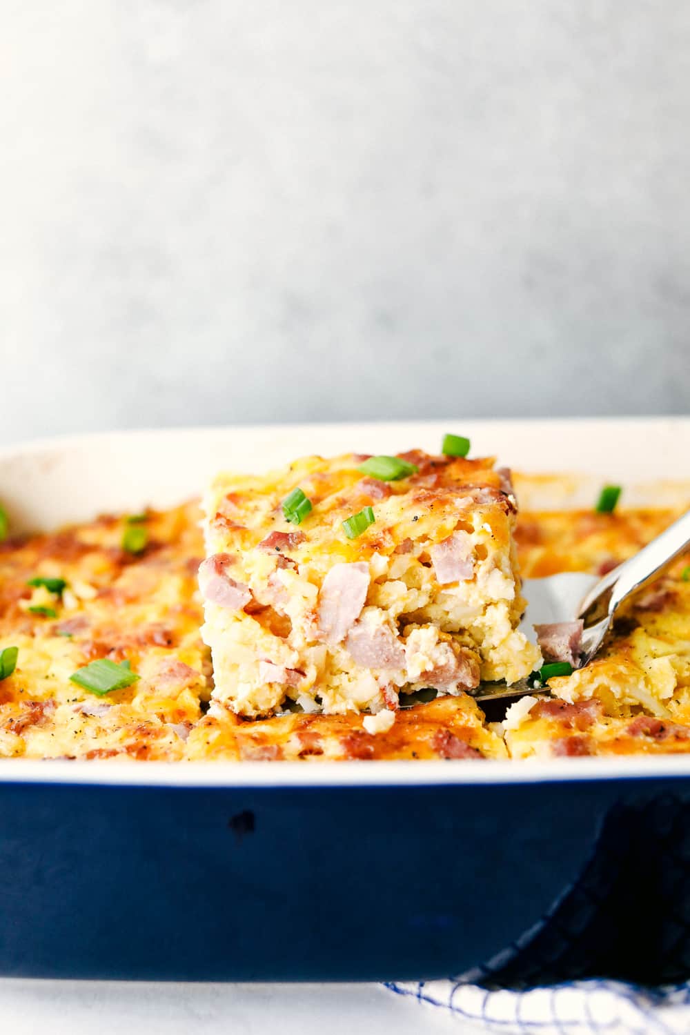 The Best Breakfast Casserole is a thick and creamy egg base with shredded potatoes, chunks of ham and flavorful cheese blends that bakes up in no time. Make ahead or enjoy the morning of during the holidays! Side shot of someone lifting out a slice of casserole.
