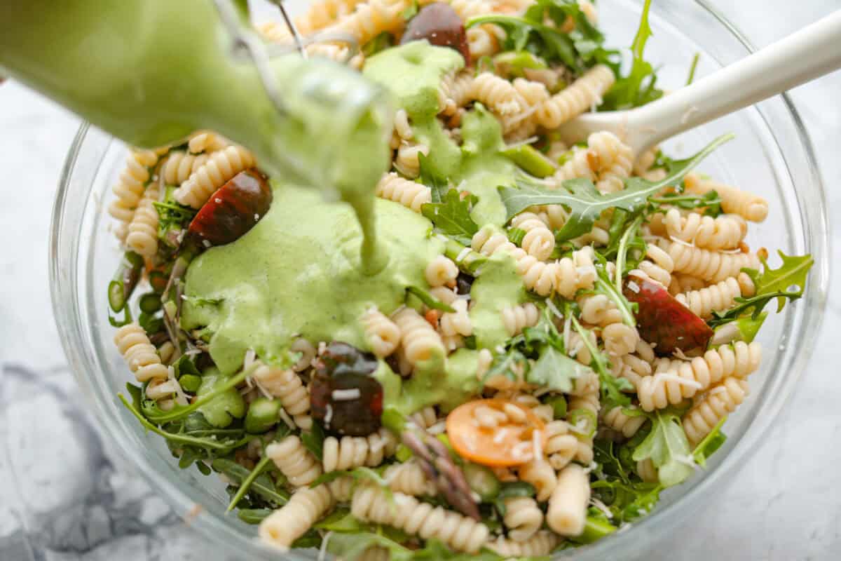 This Green Goddess Pasta Salad is fresh, vibrant, and full of flavor! The creamy herb dressing adds a tangy twist, while crisp spring veggies make it light yet hearty. Perfect for potlucks or any time you’re craving a delicious side! Process shot of someone pouring the dressing over the combined salad ingredients.