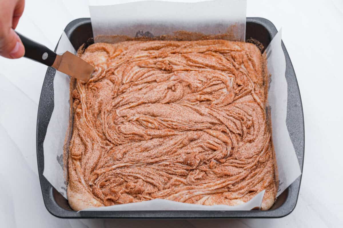 This 4-ingredient churro fudge is pure magic! It's buttery, cinnamon sugary bliss that tastes like a churro and fudge had a baby. It’s easy to make and even easier to gift (if you can resist eating it all first!). Someone swirling the cinnamon and sugar added to the top of the fudge mixture in the pan.