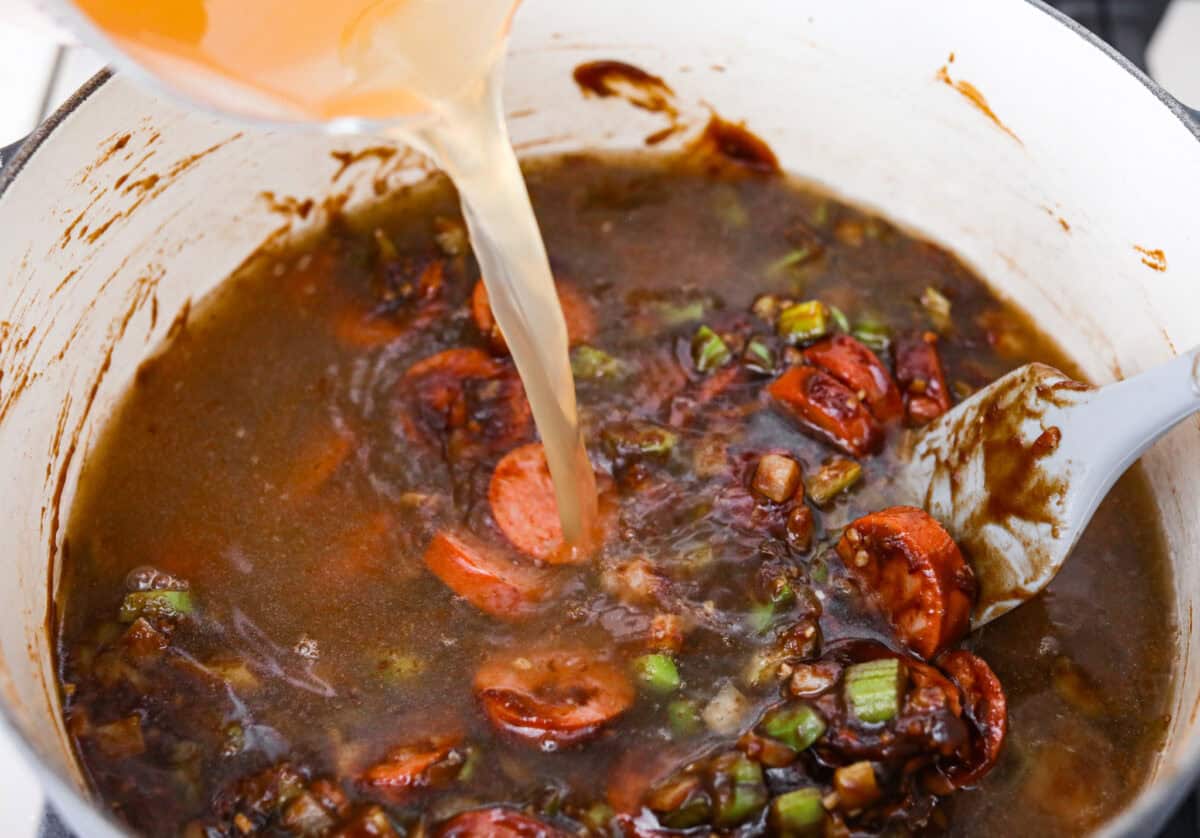 Gumbo is one of those recipes everyone makes differently, but this chicken and sausage gumbo keeps it simple. A dark roux, andouille sausage, and rotisserie chicken give you big flavor with minimal effort. Someone pouring chicken broth into the roux mixture and seasonings.