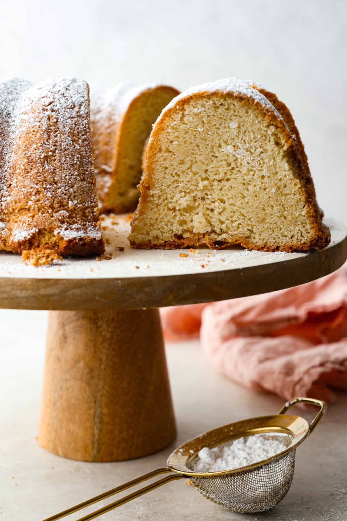 This sour cream pound cake is buttery, moist, and so tender. The light vanilla flavor with a little tang from the sour cream makes it delicious on its own, or even better with berries, whipped cream, or ice cream. Sour cream pound cake on a cake stand with a tine gold sifter with powdered sugar in it in the foreground.