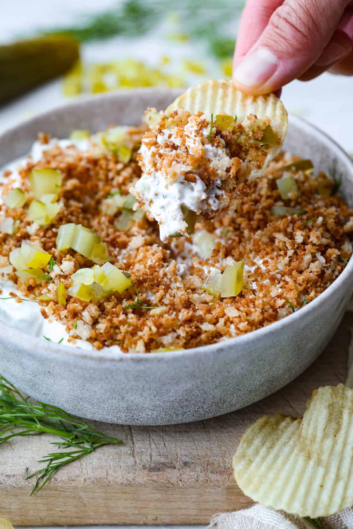 Meet my Fried Pickle Dip...a viral take on my popular pickle dip! The toasted bread crumbs elevate the flavors with the crunchy "fried" texture. Someone taking a scoop of fried pickle dip with a ruffle chip.