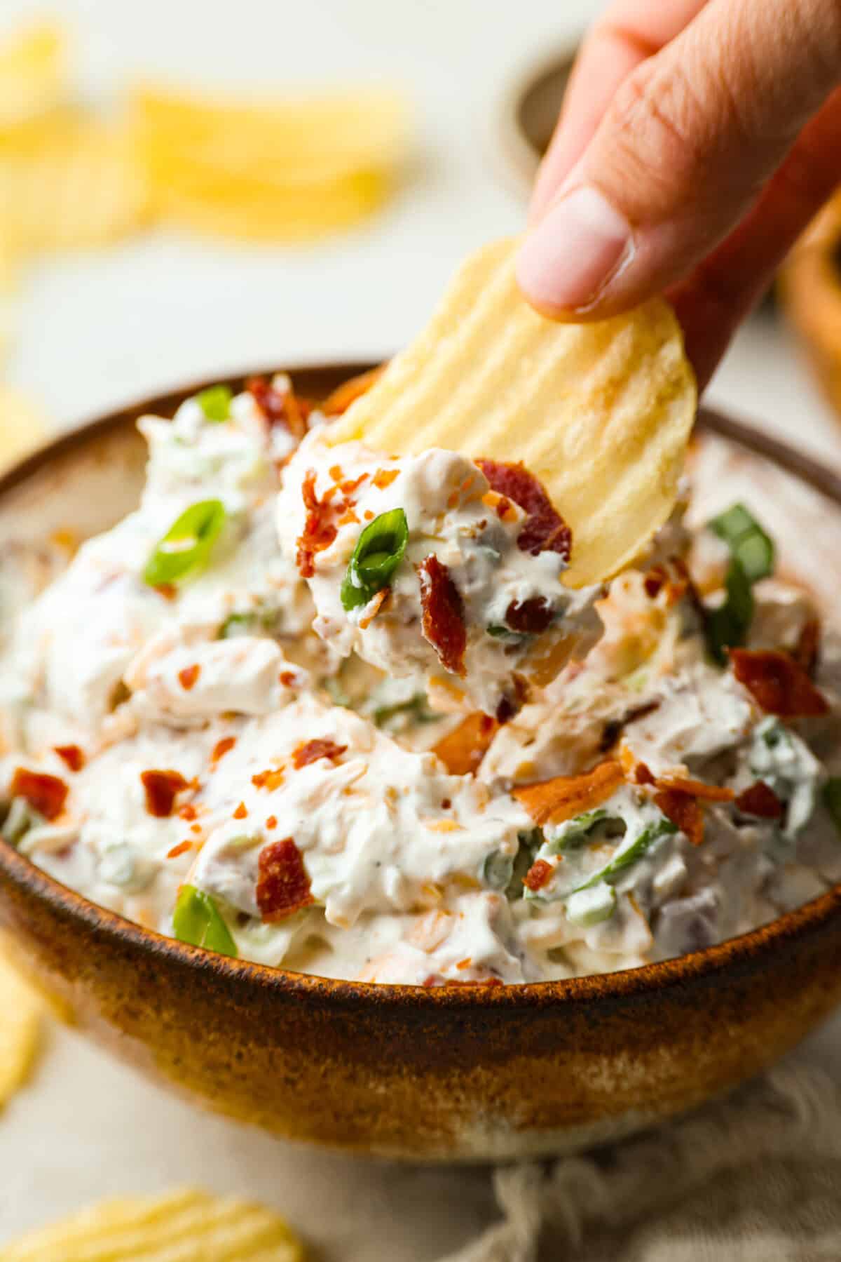You need to stop what you’re doing and make this loaded baked potato dip right now! It’s creamy, cheesy, bacony perfection! Literally to die for! It's perfect for game day, parties, or to snack on while you catch up on your favorite show. Someone using a ruffled chip to take a scoop of loaded baked potato dip.