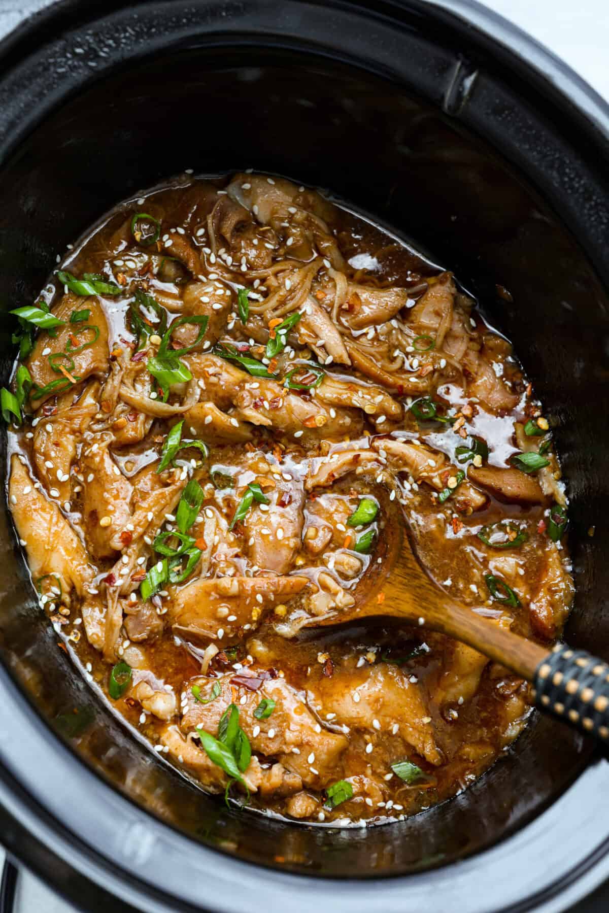 Set it and forget it! This slow cooker Korean BBQ chicken is sweet, savory, and just the right amount of spice. Perfect for tossing over rice, piling onto buns, topping a salad, or even making tacos. Weeknight dinner just got easier! Slow cooker Korean BBQ chicken in a crockpot with a wooden spoon, with sesame seeds and green onions.