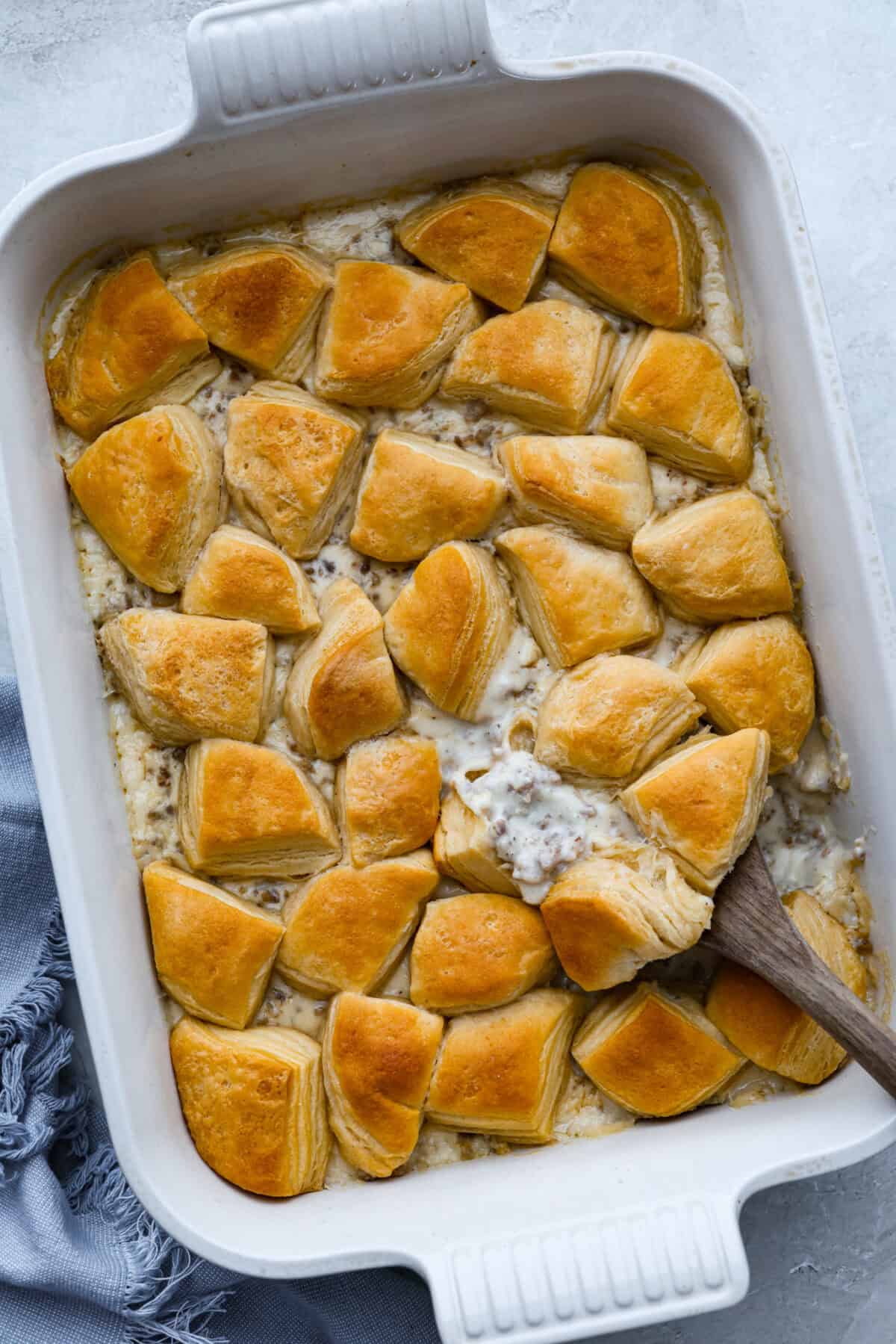 Forget the usual biscuits and gravy, this version takes everything you love and turns it into one irresistible, bubbly casserole. Creamy sausage gravy, buttery biscuits, and zero stress. Casserole dish with biscuits and gravy casserole baked to perfection and a wooden spoon taking a scoop out.