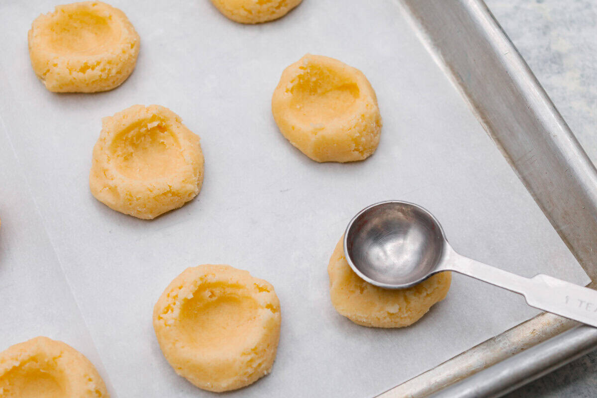 Meet the upgraded Twix thumbprint cookies! I re‑tested and updated the recipe so you'll get defined wells, clean edges, and a tender center every time! They are perfect for gift giving and cookie parties! Someone pressing the bottom of a rounded teaspoon into the top of the cookie dough ball before its baked.