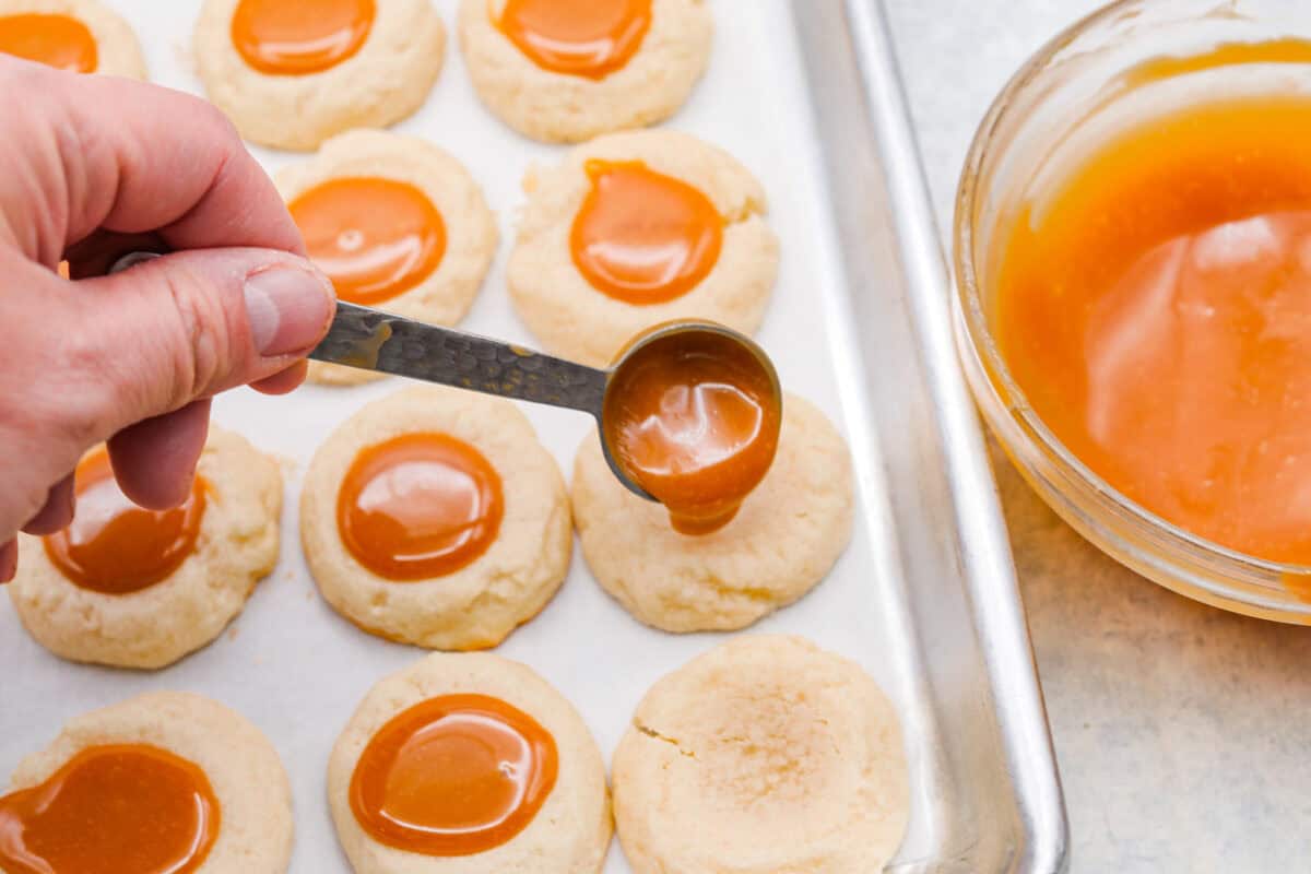 Meet the upgraded Twix thumbprint cookies! I re‑tested and updated the recipe so you'll get defined wells, clean edges, and a tender center every time! They are perfect for gift giving and cookie parties! Someone spooning a small amount of caramel into the thumbprint well in the cookie.