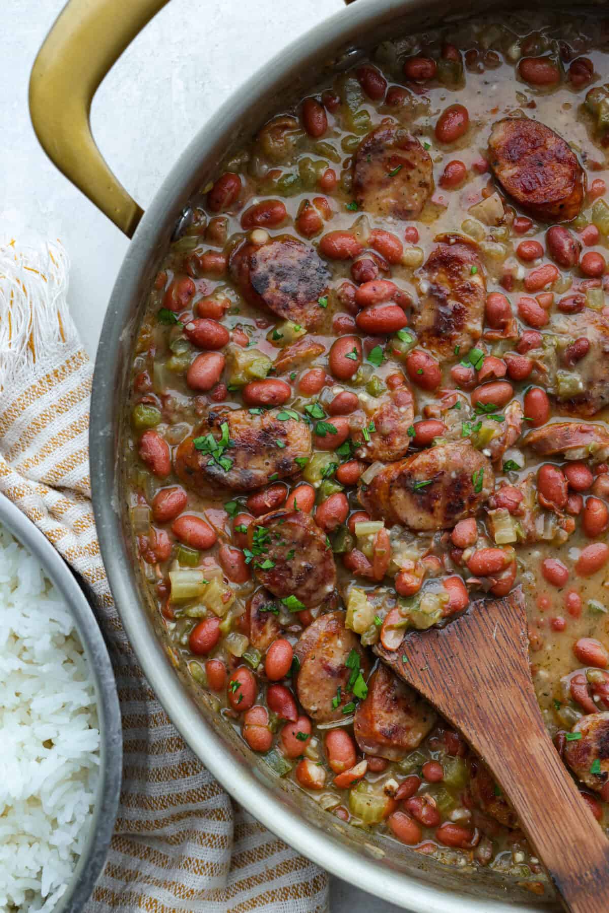 If your family is hungry now and you don't have time to soak beans or babysit the stove, this red beans and rice is for you. It is quick, cozy, and made with canned beans and smoky sausage. It tastes great and is ready fast! Skillet of red beans and rice with a wooden spoon.