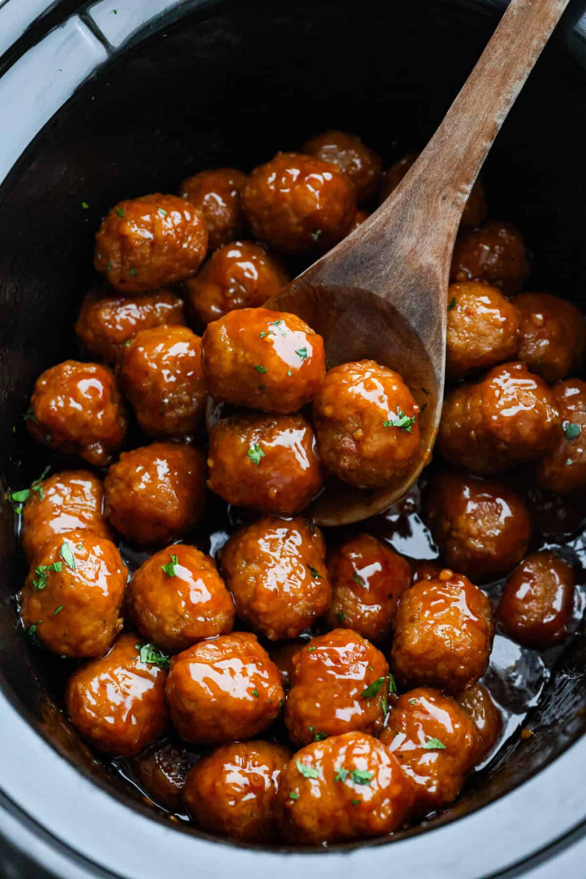 Slow cooker bourbon meatballs are an easy, set-it-and-forget-it favorite. Frozen meatballs simmer in a sweet, sticky bourbon sauce, making them perfect for an appetizer or a simple dinner with just 10 minutes of prep. Someone taking a large wooden spoon and scooping a few slow cooker bourbon meatballs out of the crockpot.