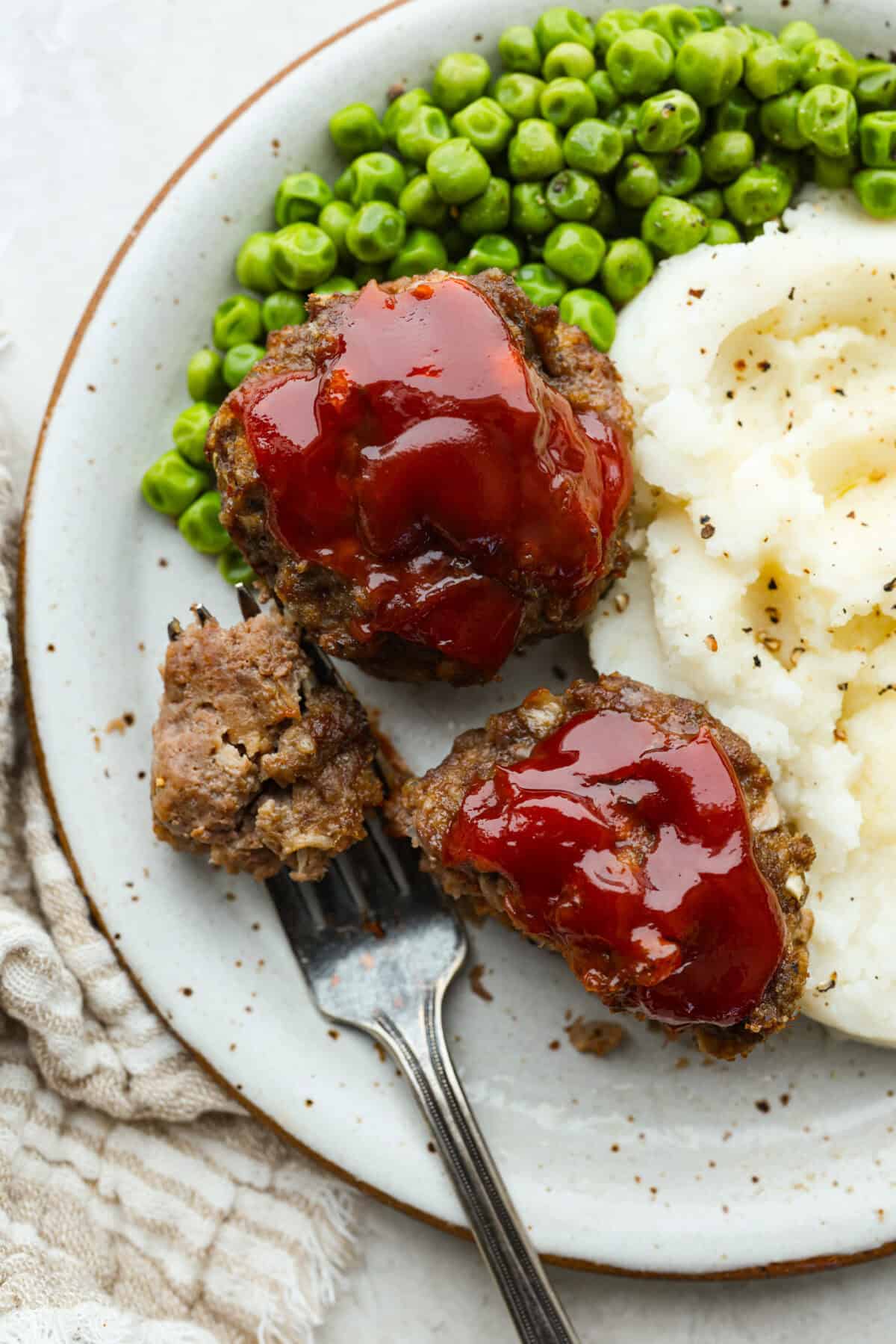 Plated mini meatloaf muffins next to mashed potatoes and peas. 